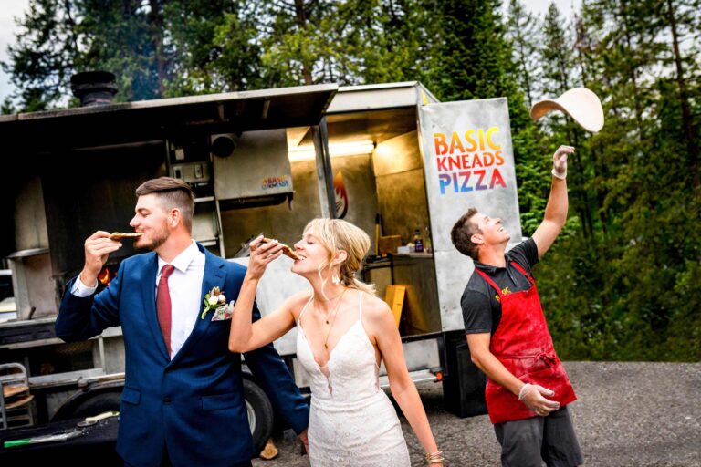 A bride and groom pose eating pizza while a food truck vendor tosses a crust in the air behind them