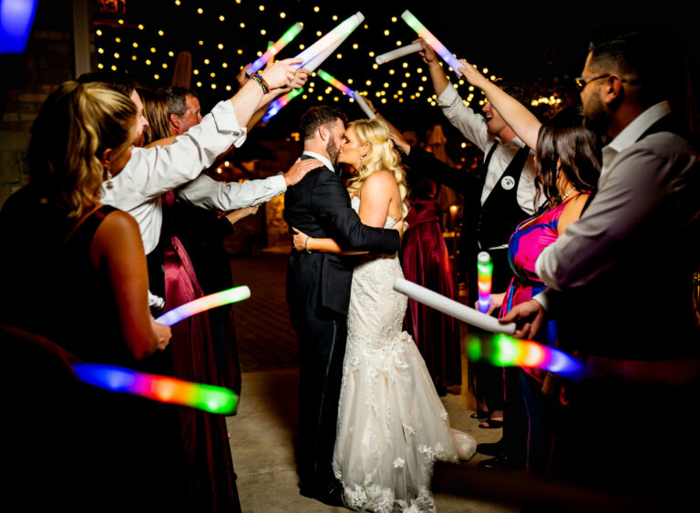 A bride and groom embrace surrounded by their guests waving glow sticks and photographed in a candid documentary wedding photography style at Della Terra Mountain Chateau in Estes Park