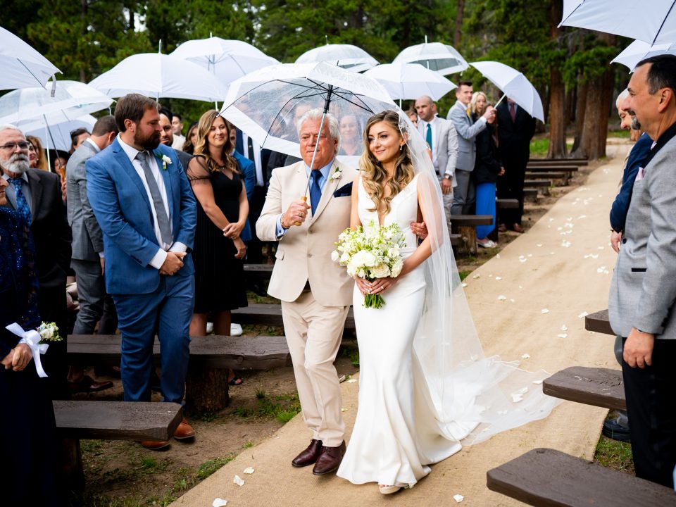 A bride is walked down the aisle by her father at the Dao House as guests stand with umbrellas