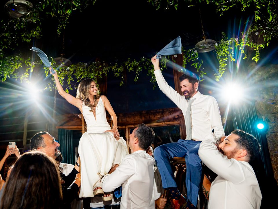 A bride and groom wave napkins above their heads as they're lifted into the air on chairs in the Dao House's reception all