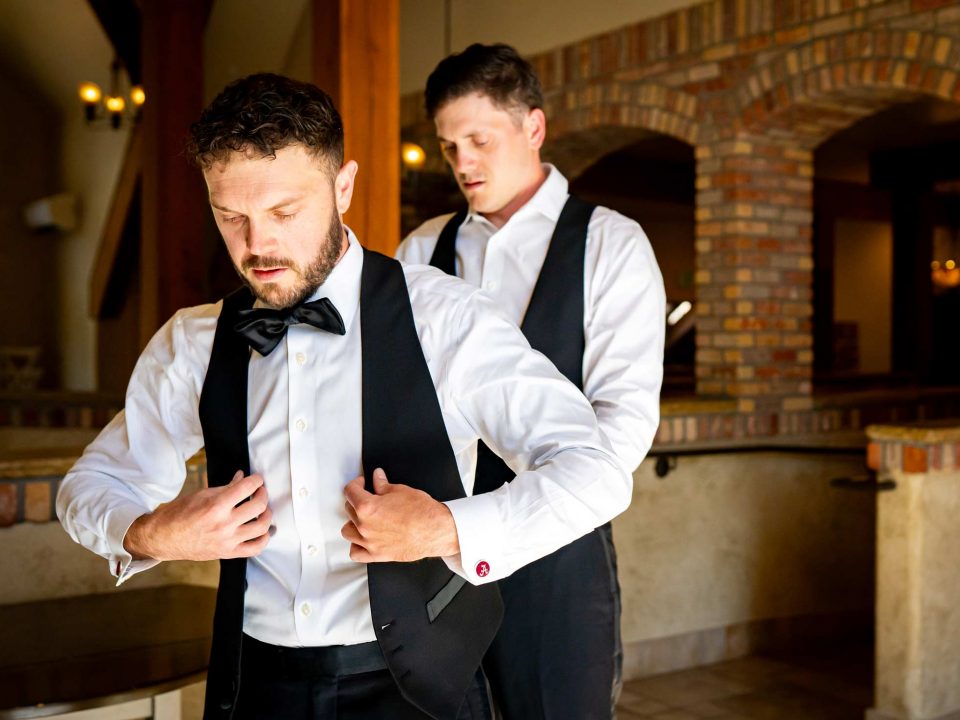 A groom's brother helps him get his vest on while getting ready for his Della Terra Mountain Chateau wedding