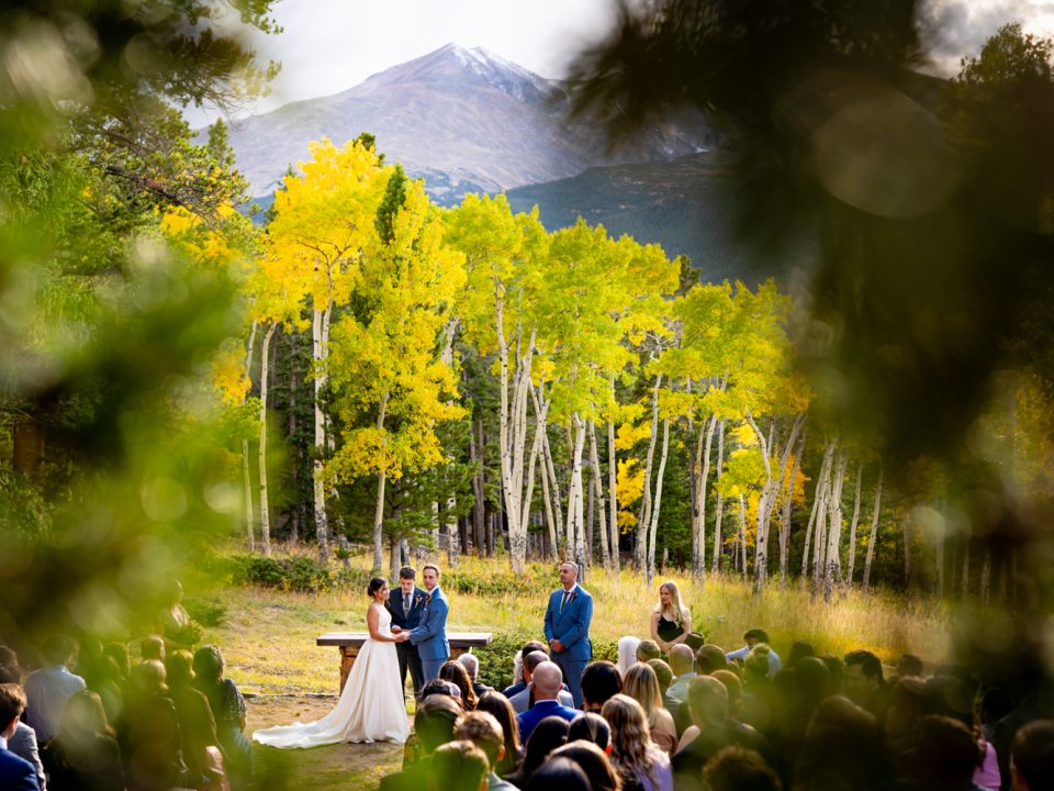 A bride and groom stand together during the ceremony of their Dao House wedding with Longs Peak and golden aspens in the background